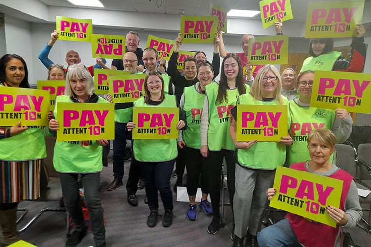 Group of teachers in green tabards holding up signs saying "Pay attention' with the io in attention made up of the 10 that echoes the 10 percent pay demand