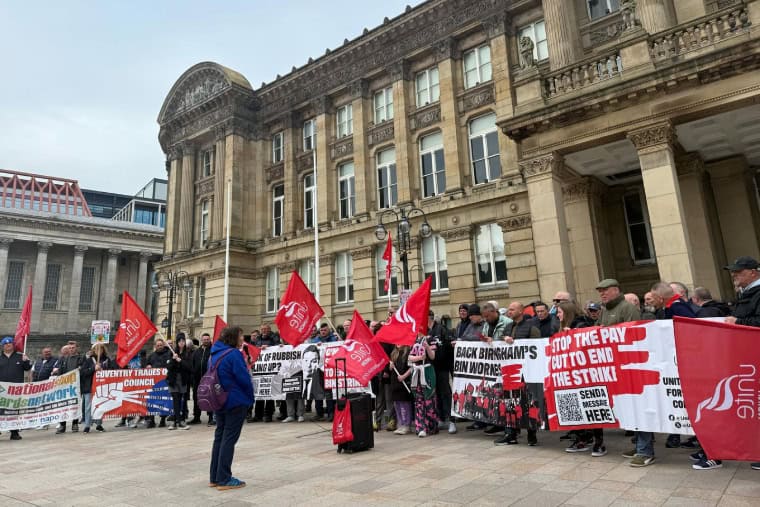 Large rally with dozens of workers with red flags being addressed by a person with sound system