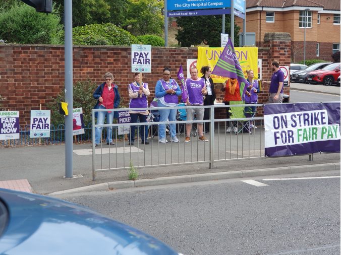 A row of workers are on a picket line. They have a number of colourful banners and there is a Unison union sign.