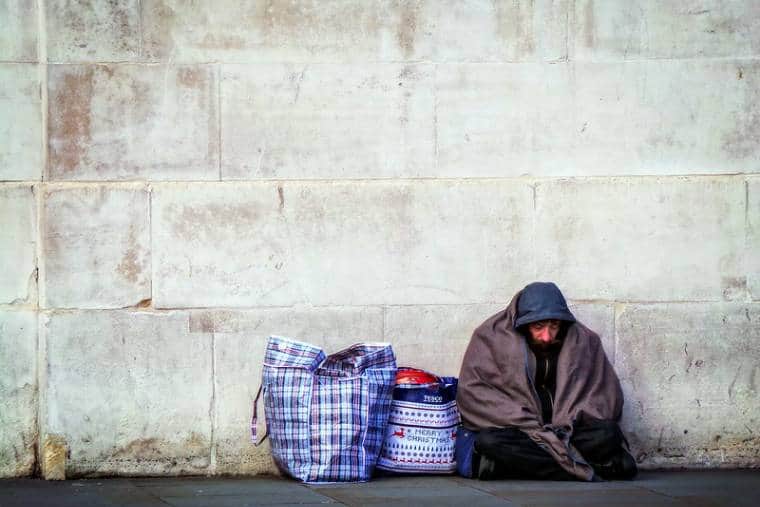 A homeless man sits against a wall beside two carrier bags of his possessions